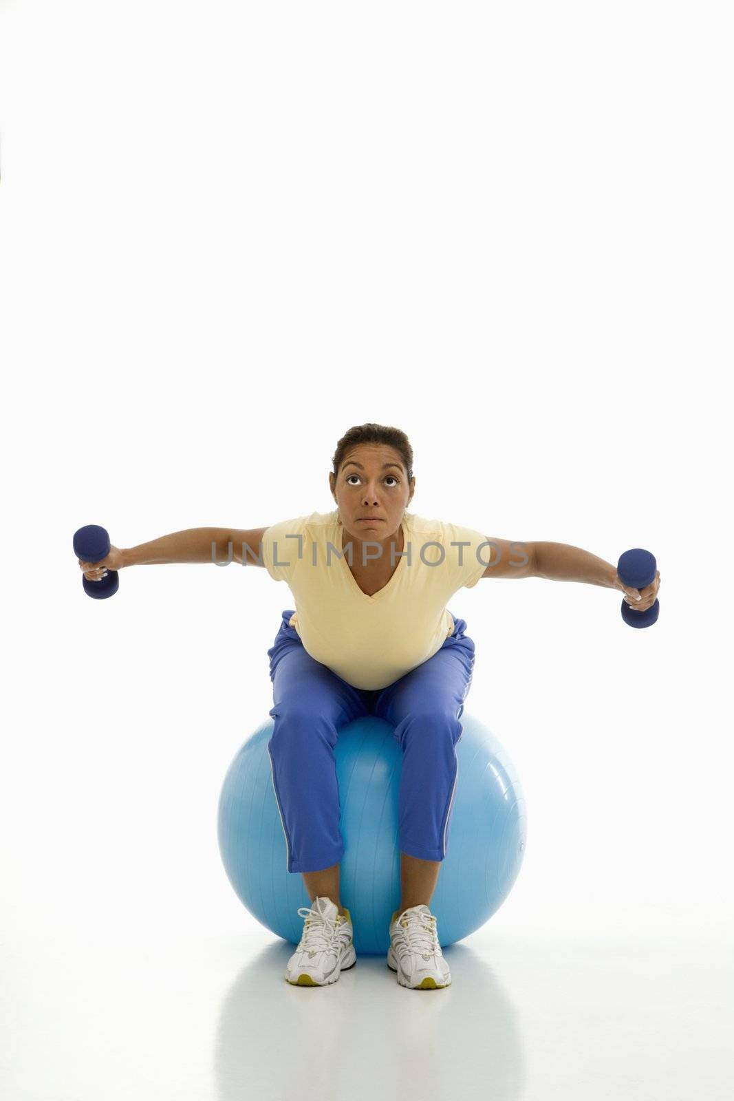 Mid adult multiethnic woman balancing on blue exercise ball with outstretched arms holding dumbbells.