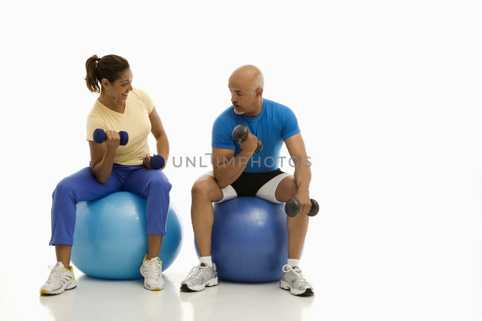 Mid adult multiethnic man and woman balancing on blue exercise balls while working out with dumbbells.