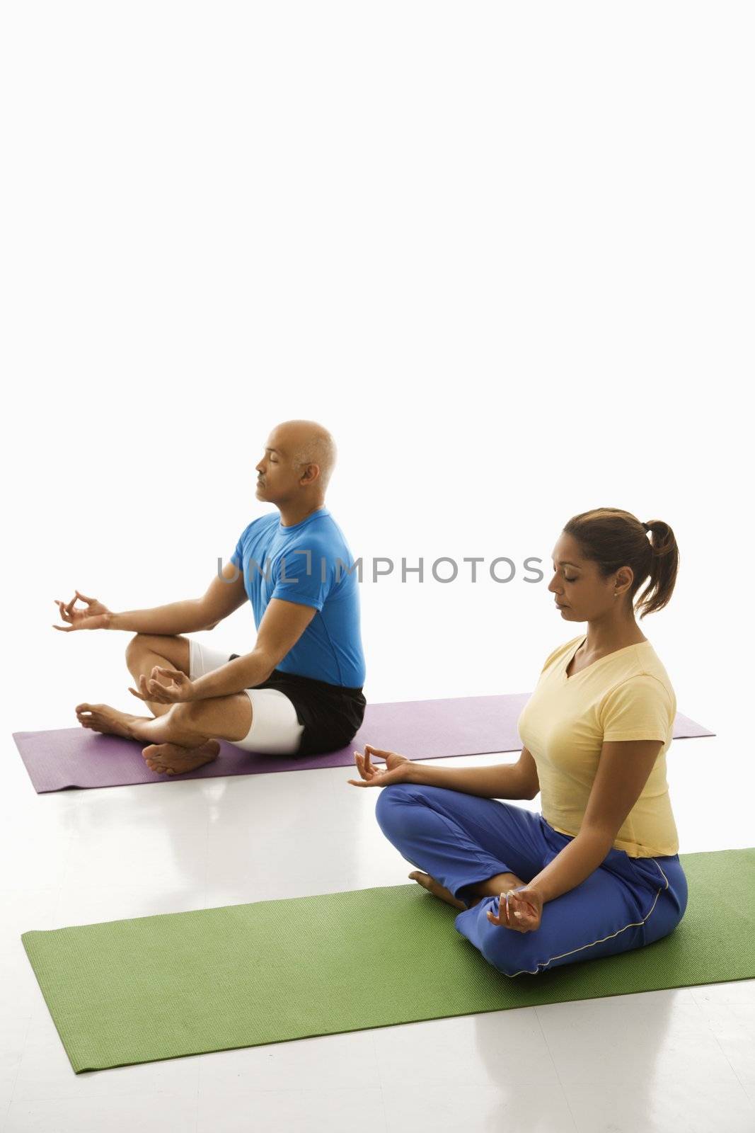 Side view of mid adult multiethnic man and woman sitting in lotus position on exercise mats with eyes closed and legs crossed.