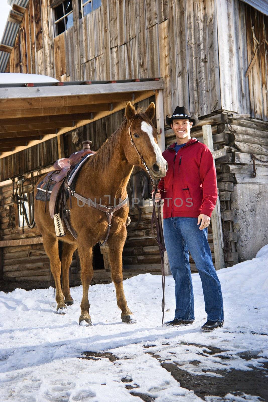 Young Caucasian man in western attire holding his horse with stable in background.