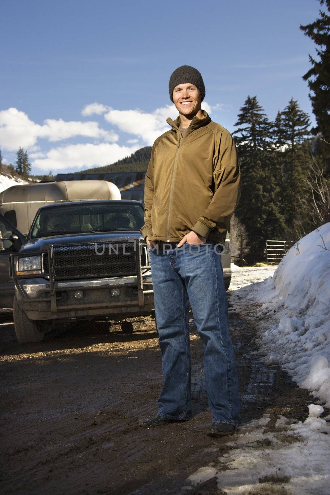 Young Caucasian man standing in front of truck with hands in pockets smiling at viewer.