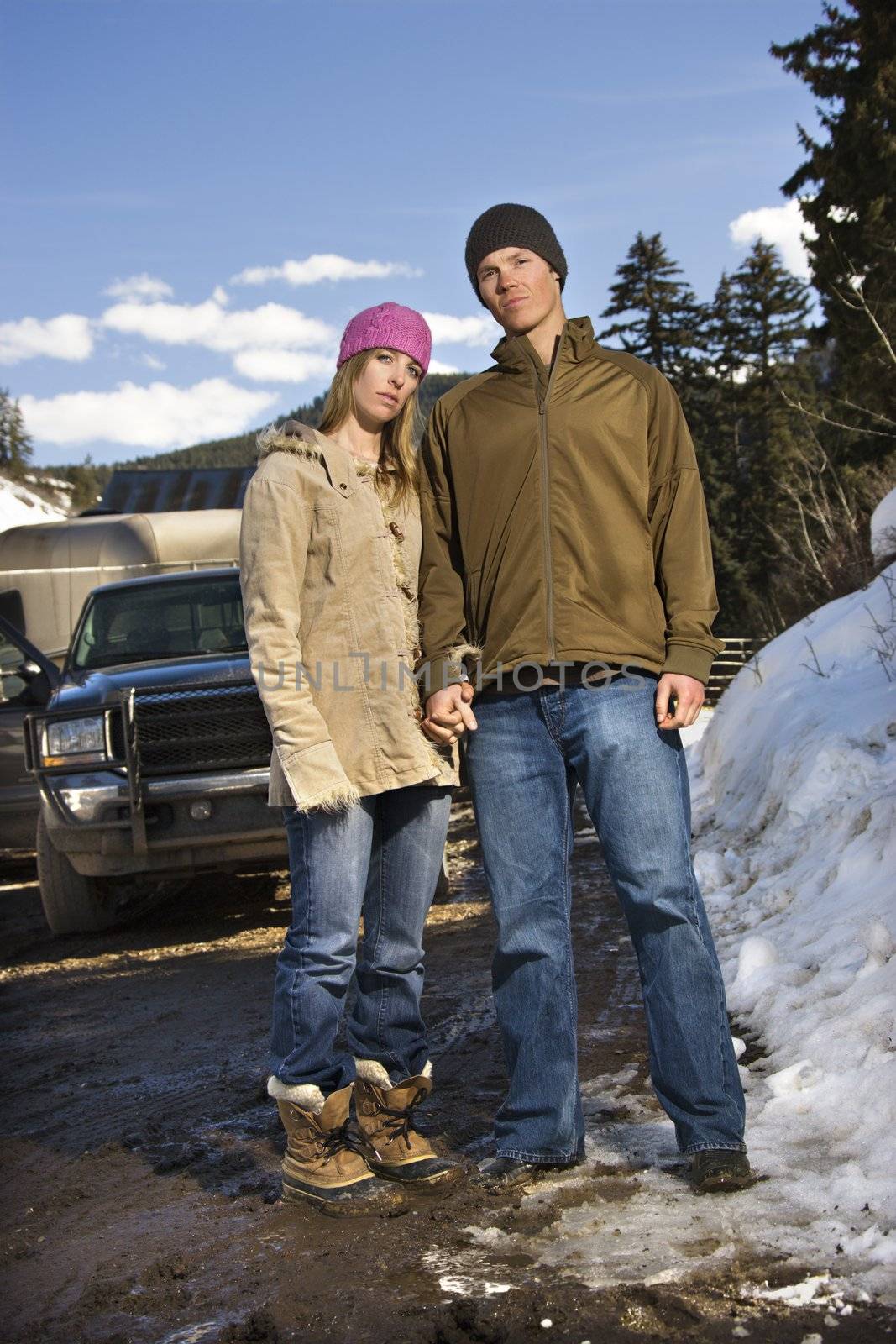Young Caucasian couple holding hands looking at viewer with truck in background.