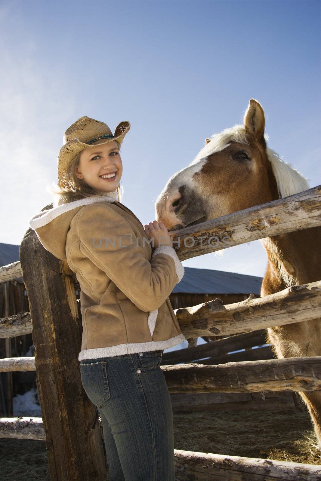 Young Caucasian woman standing at fence with horse looking at viewer.