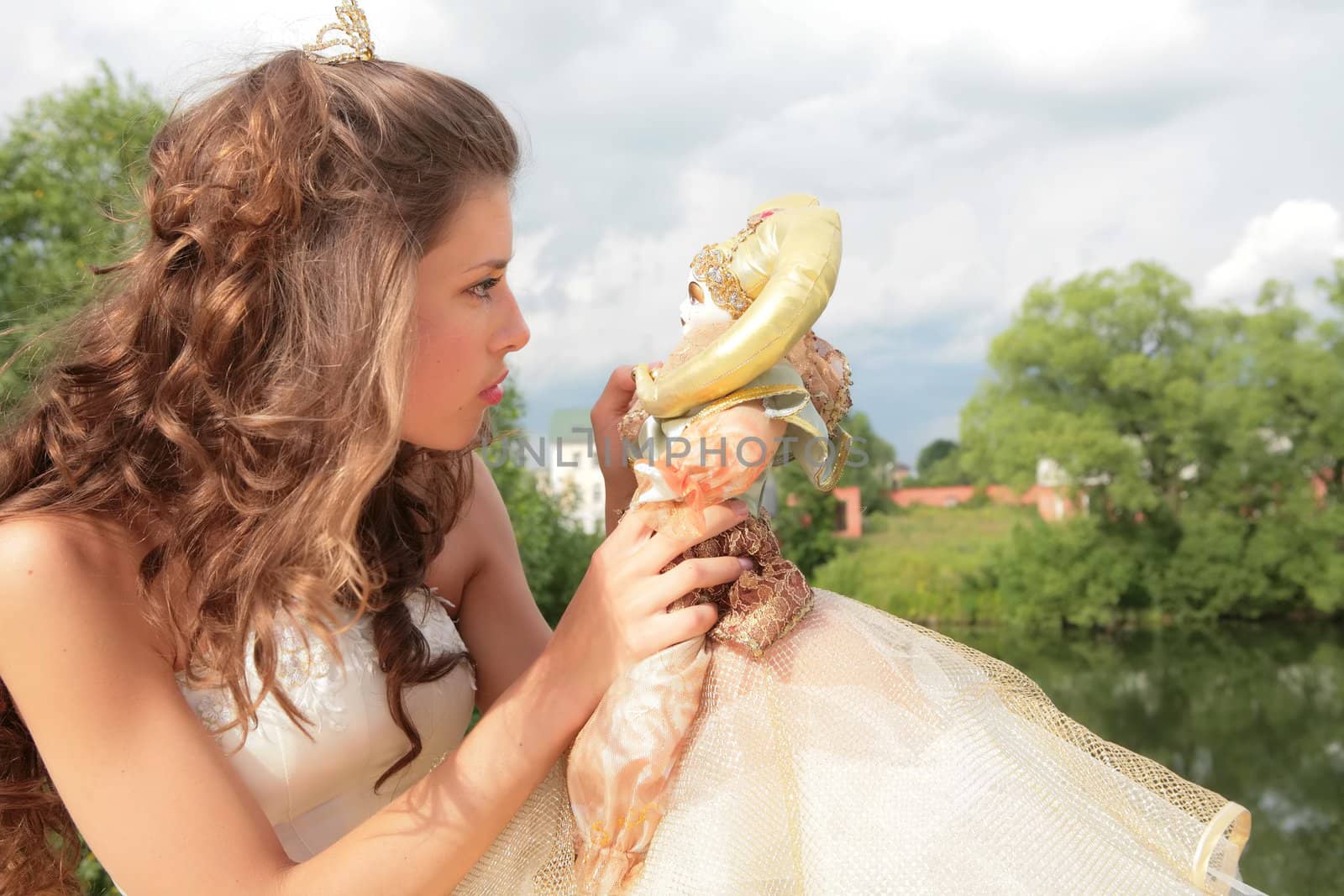 princess in white-golden gown plays with loved doll on background cloudy sky