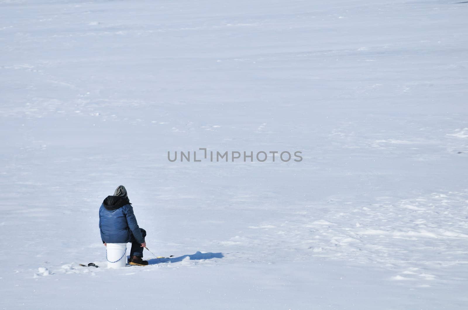 Ice fisherman sitting on a bucket above hole in the ice.