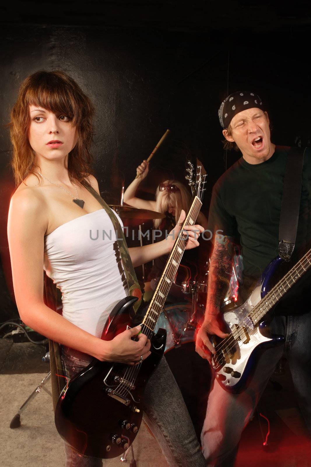 Band playing on a stage. Female guitarist, male bassist and female drummer. Shot with strobes and slow shutter speed to create lighting atmosphere and blur effects. Slight motion blur on performers.
