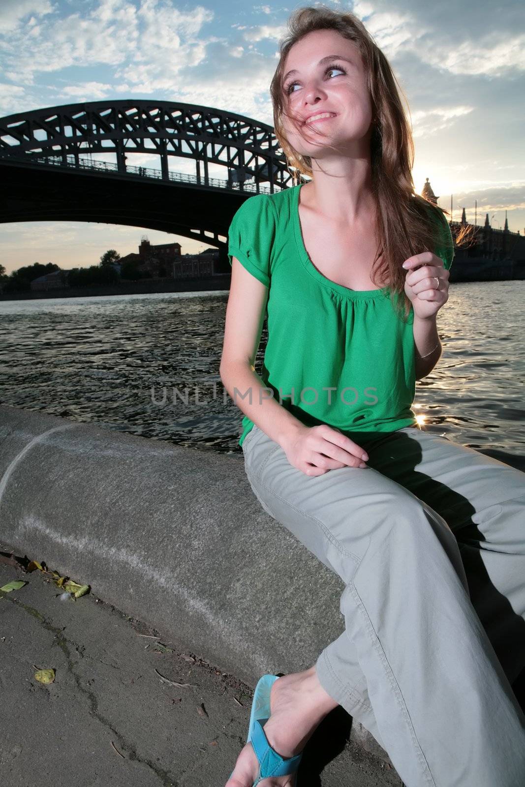 smiling girl sits on granite quay Moscow-river on background of the bridge, water and sky