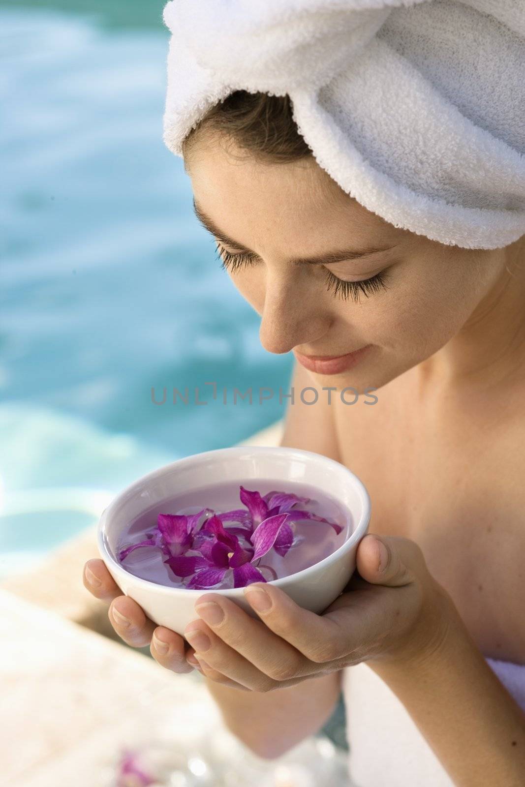 Caucasian mid-adult woman wearing towel around head and body holding bowl of purple orchids next to pool.