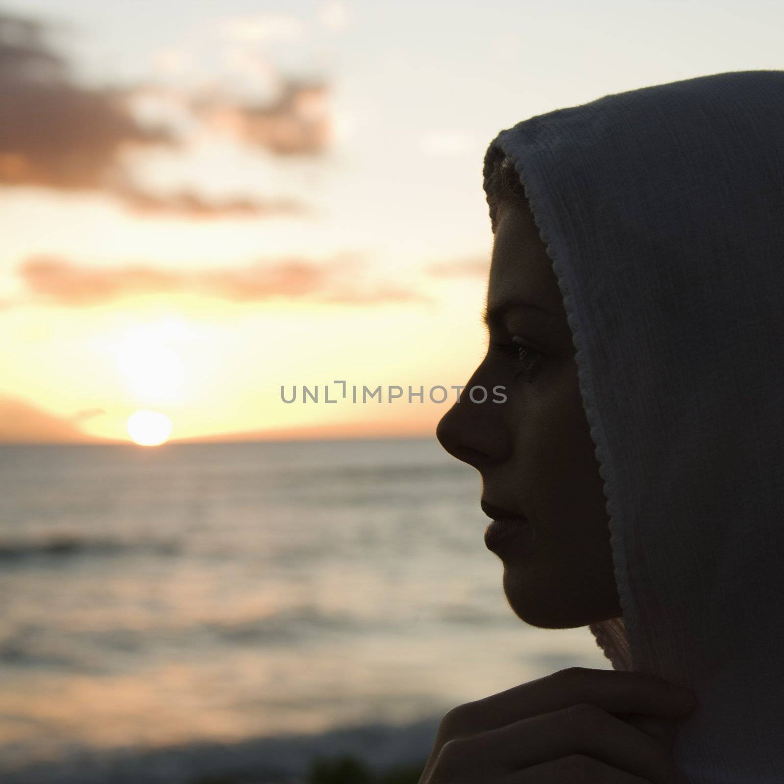 Pretty Caucasian mid-adult woman with scarf over head profiled against sunset over ocean.