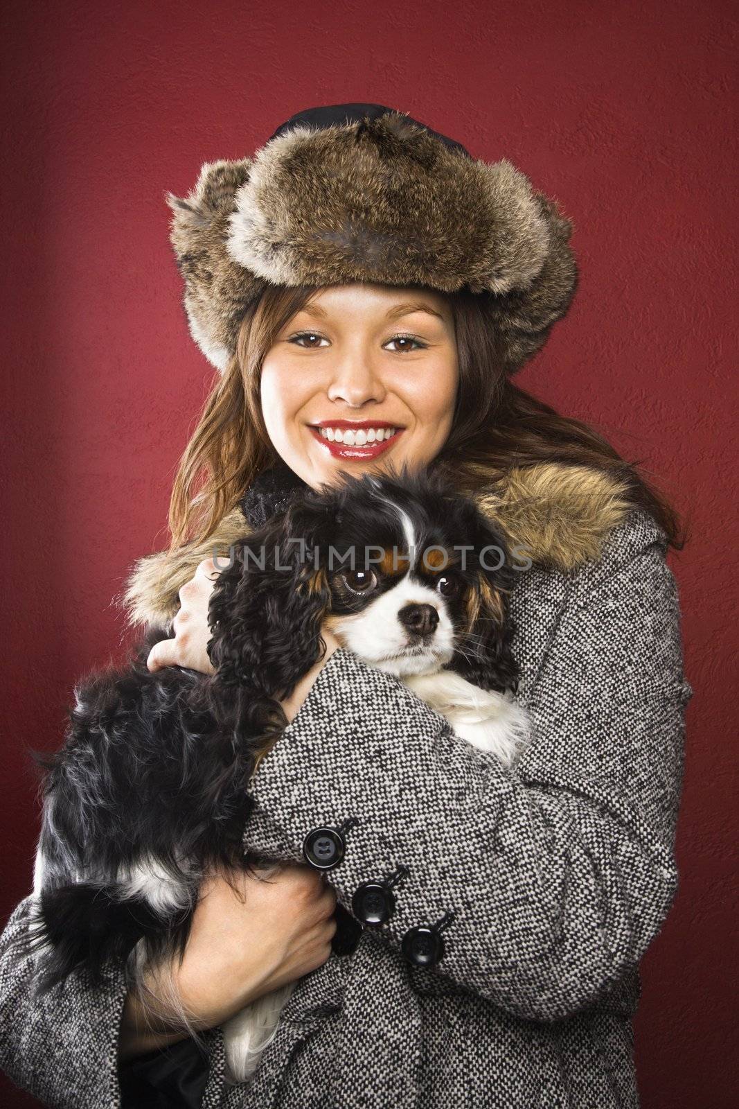 Young adult Caucasian woman wearing fur hat holding King Charles Spaniel in arms and smiling at viewer.