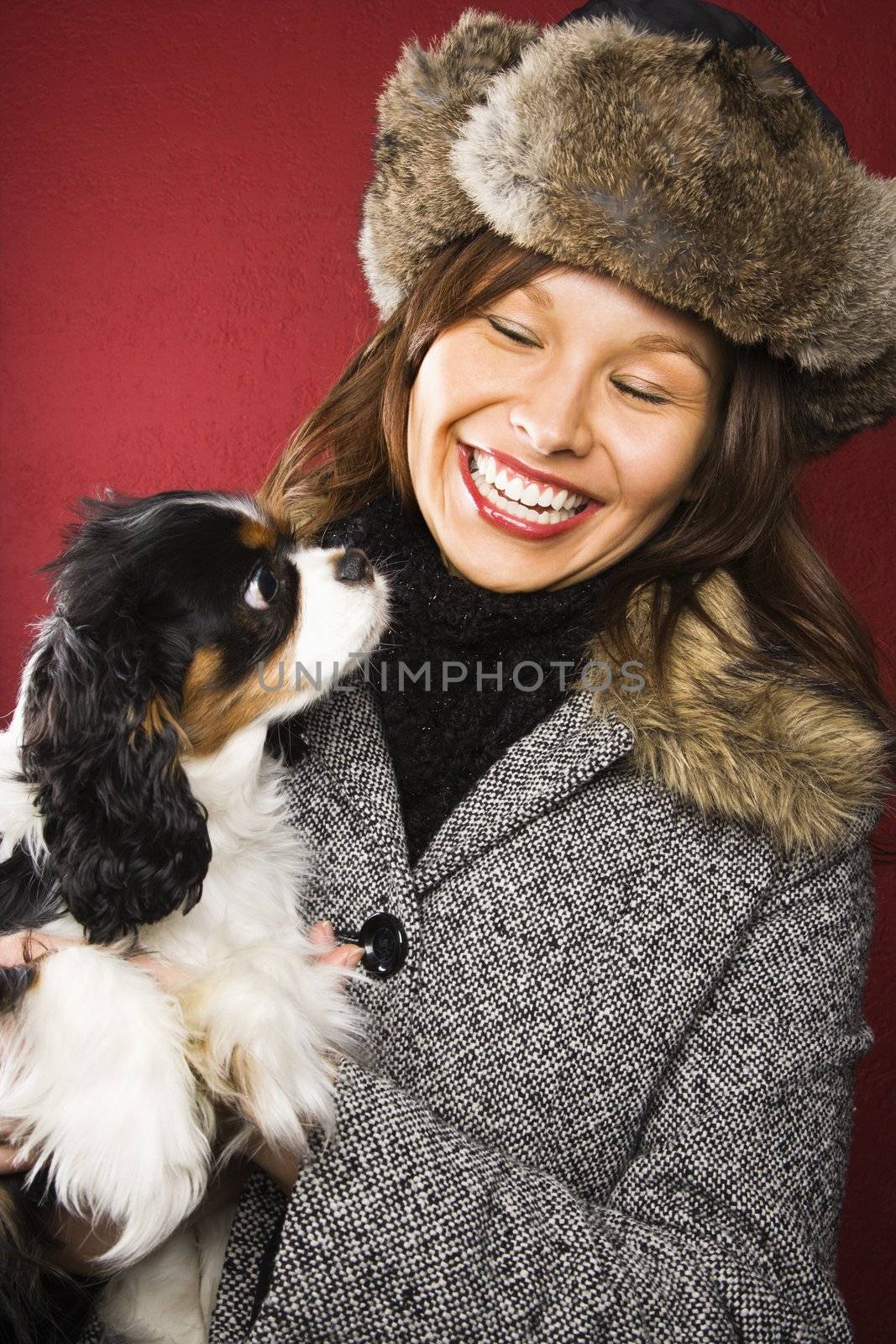 Young adult Caucasian woman wearing fur hat holding King Charles Spaniel in arms and smiling.