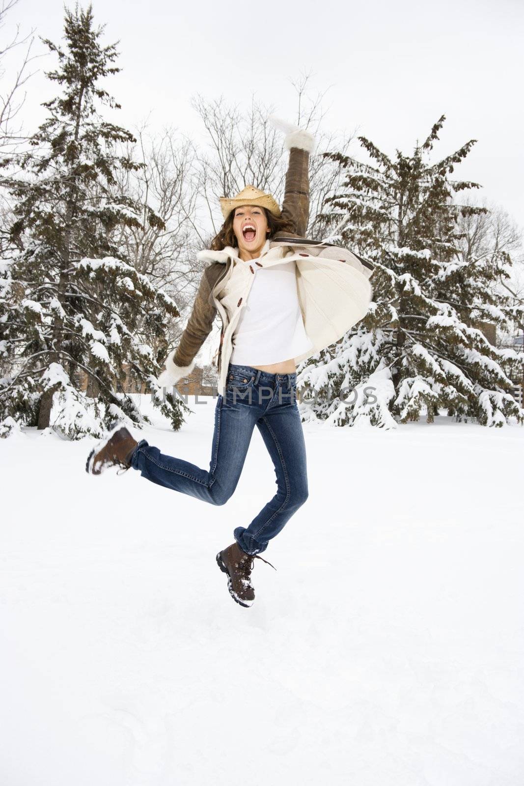 Caucasian young adult female leaping into the air in snowly climate wearing straw cowboy hat.