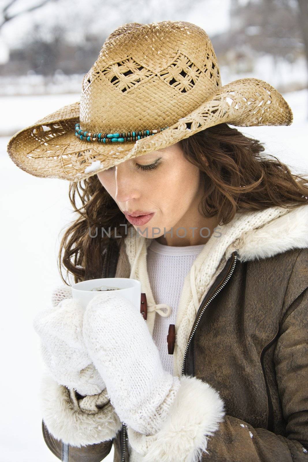 Caucasian young adult female outdoors in snow blowing into coffee cup and wearing straw cowboy hat.