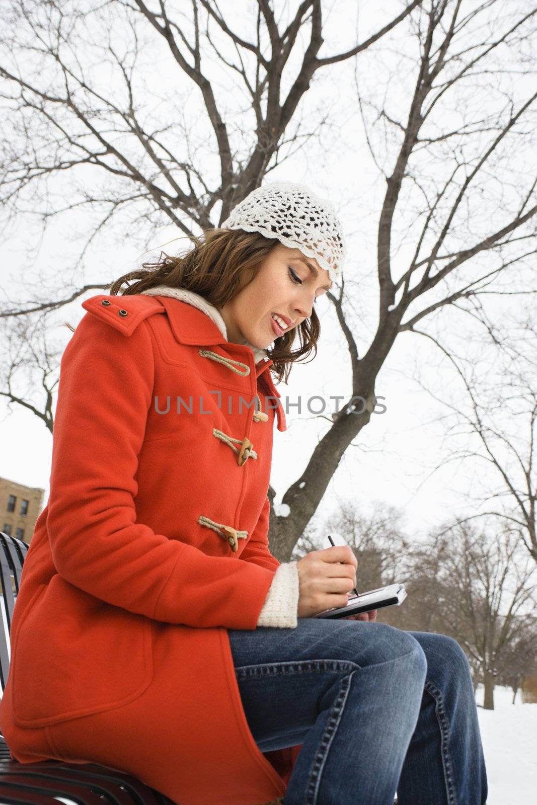 Low angle view of Caucasian young adult female in winter clothing sitting on park bench using PDA .