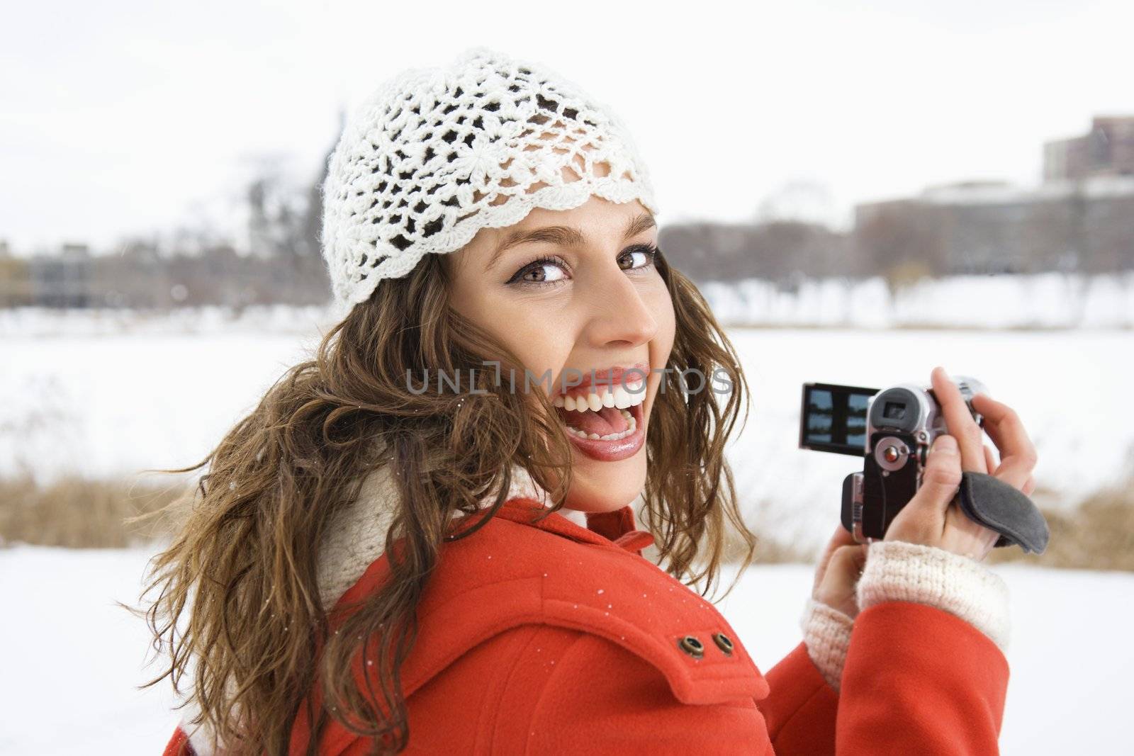 Smiling Caucasian young adult female in winter clothing holding video camera and looking over shoulder at viewer.