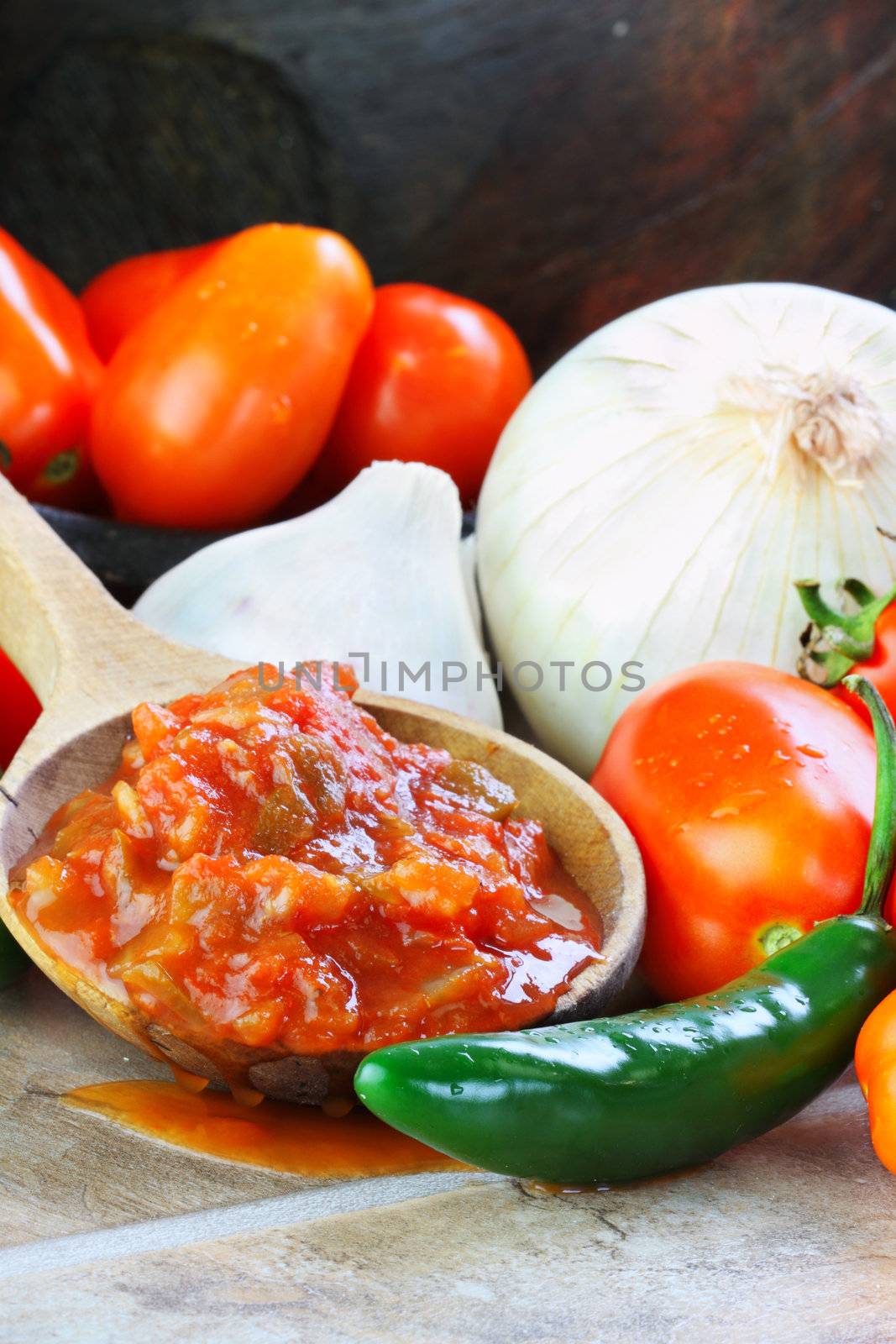 Rustic wooden spoon filled with spicy fresh salsa, surrounded by freshly picked and washed ingredients spilling from an old wooden bowl.