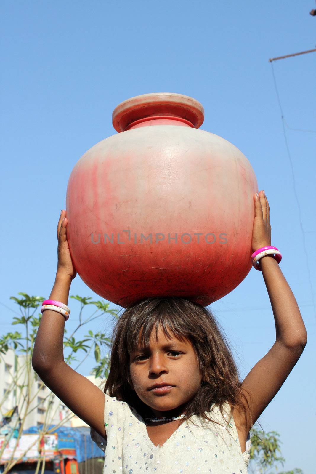 A poor Indian girl carrying water to her home in a red plastic pot, due to scarcity of drinking water to poor people in India.