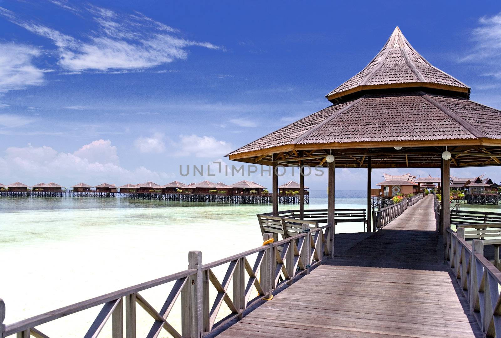 Image of a walkway and huts on stilts on a remote Malaysian tropical island with deep blue skies and crystal clear waters.