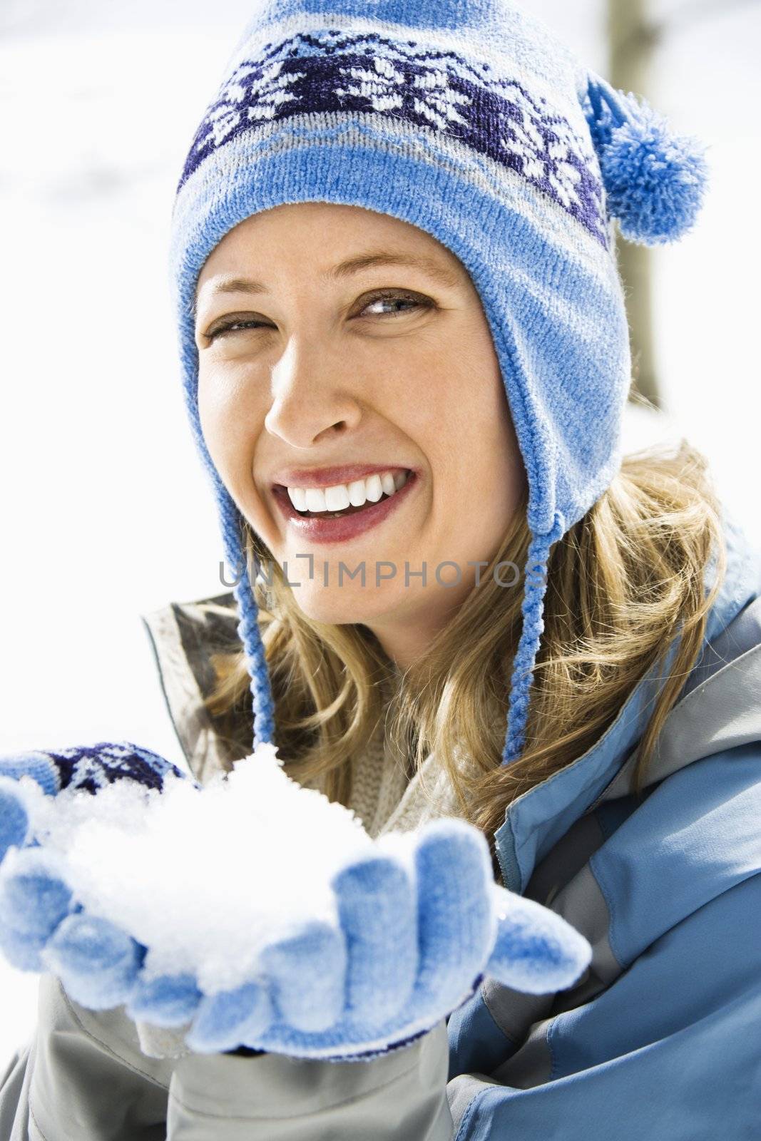 Portrait of attractive smiling mid adult Caucasian blond woman wearing blue ski cap and gloves holding snow towards viewer.