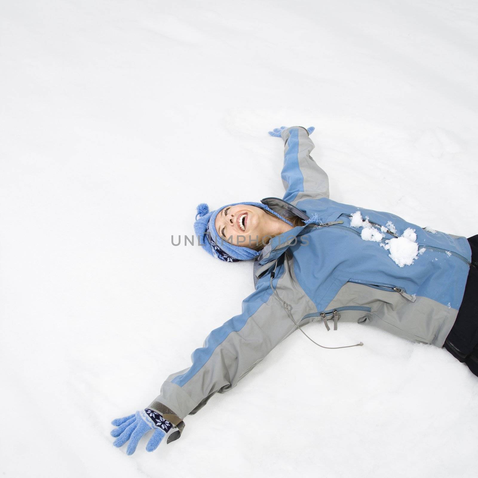 Attractive laughing mid adult Caucasian woman wearing blue ski clothing lying in snow making snow angel.