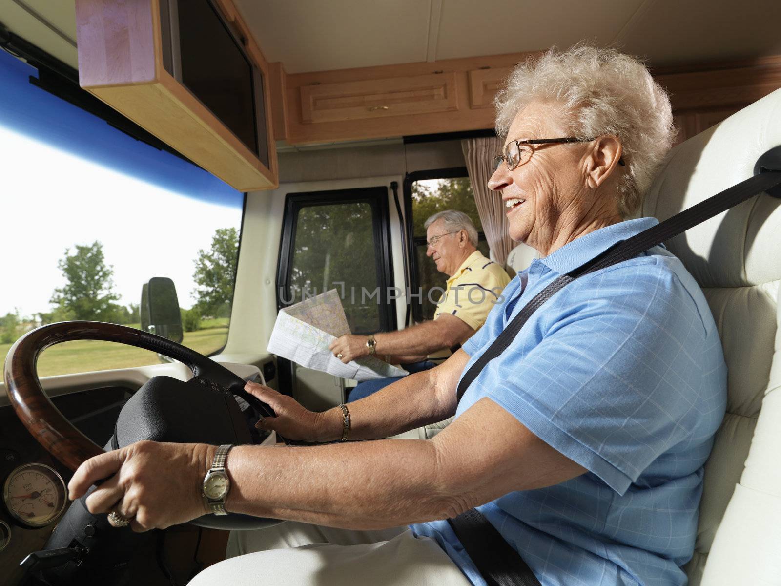 Senior adult woman driving RV and smiling while man reads map in passenger seat.