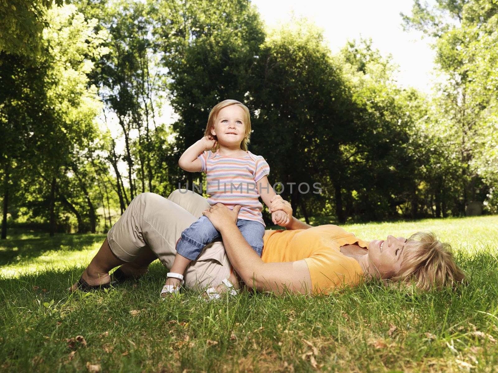 Caucasian mid adult woman lying in grass at park with toddler daughter seated on her lap.