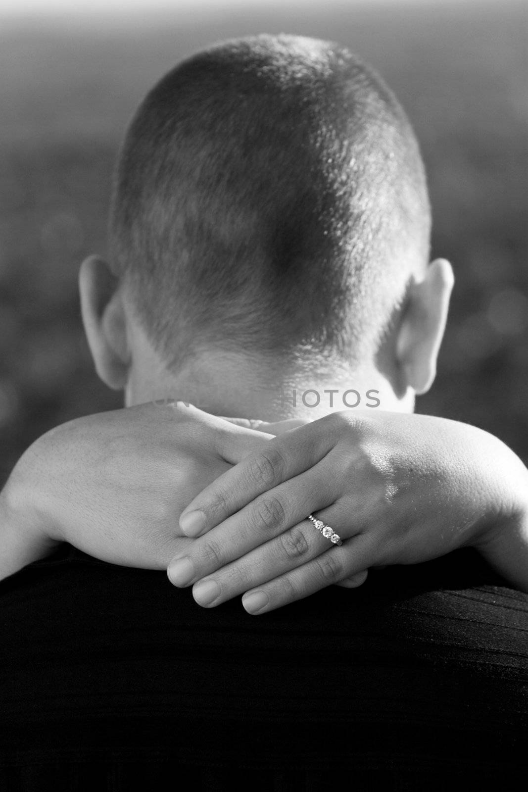 A young happy couple that is newly engaged with the woman placing her hands around her fionces neck.  Shallow depth of field with focus on the diamond engagement ring.