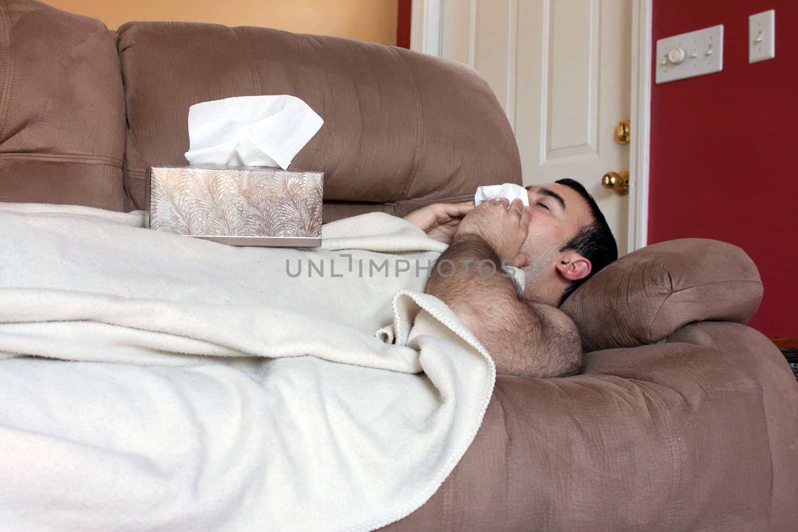 A young adult sick on the couch at home blows his nose with a tissue.