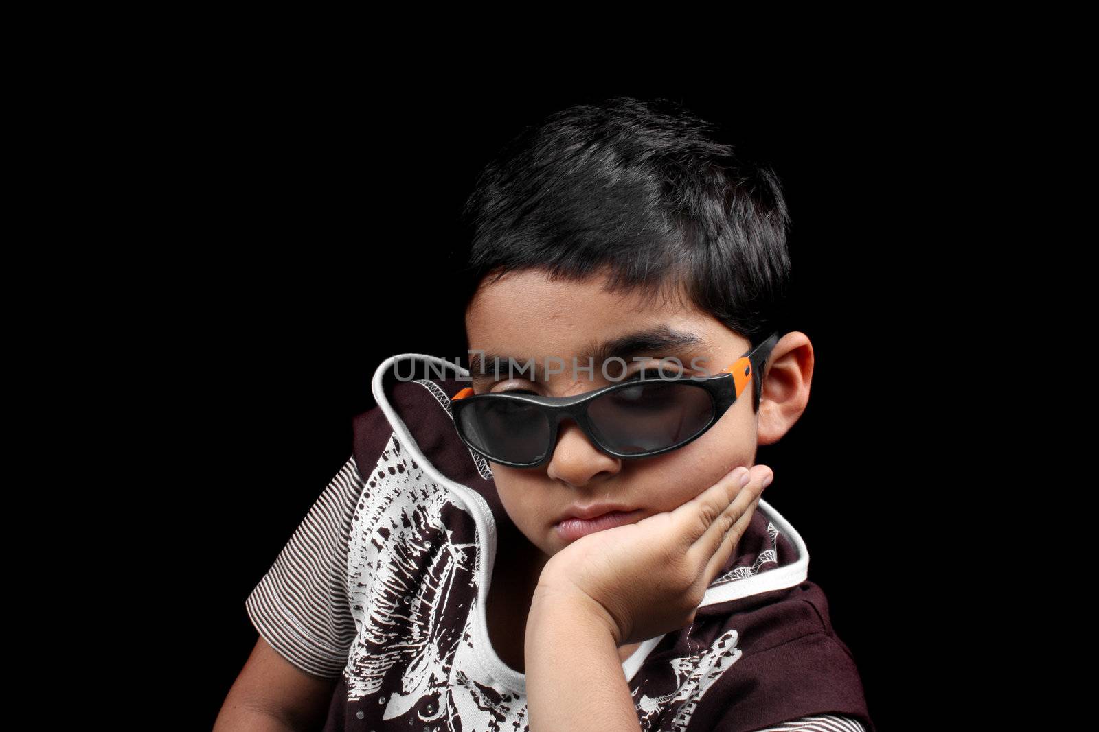 A portrait of a stylish Indian kid wearing sunglasses, on black studio background.