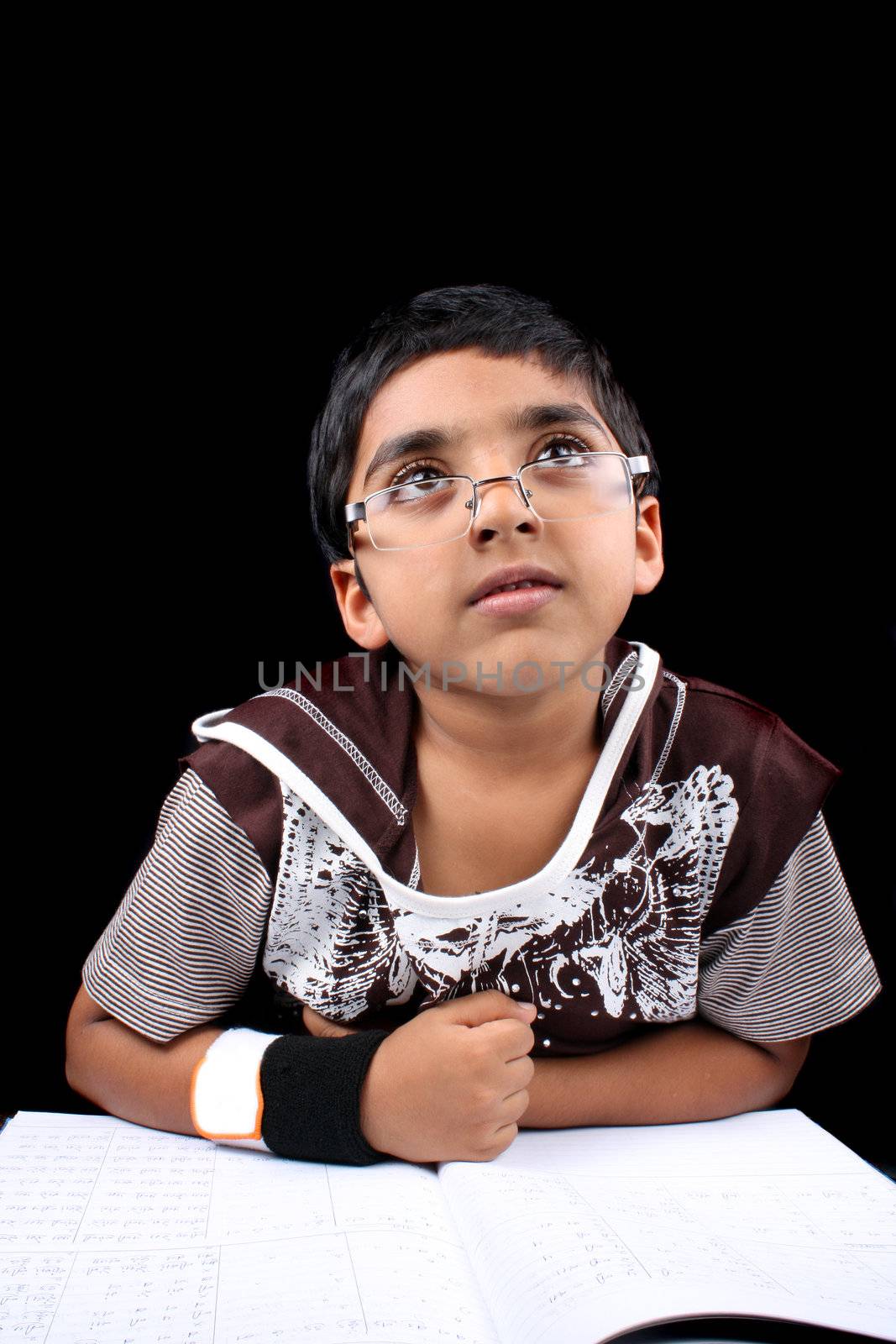 A little Indian boy lost in his dreams while doing his homework studies, on black studio background.