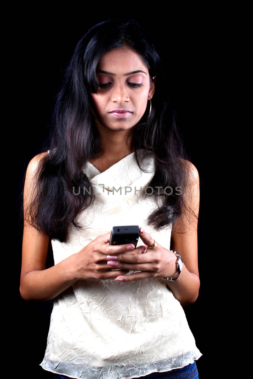A beautiful Indian teen sending a text message, on black studio background.