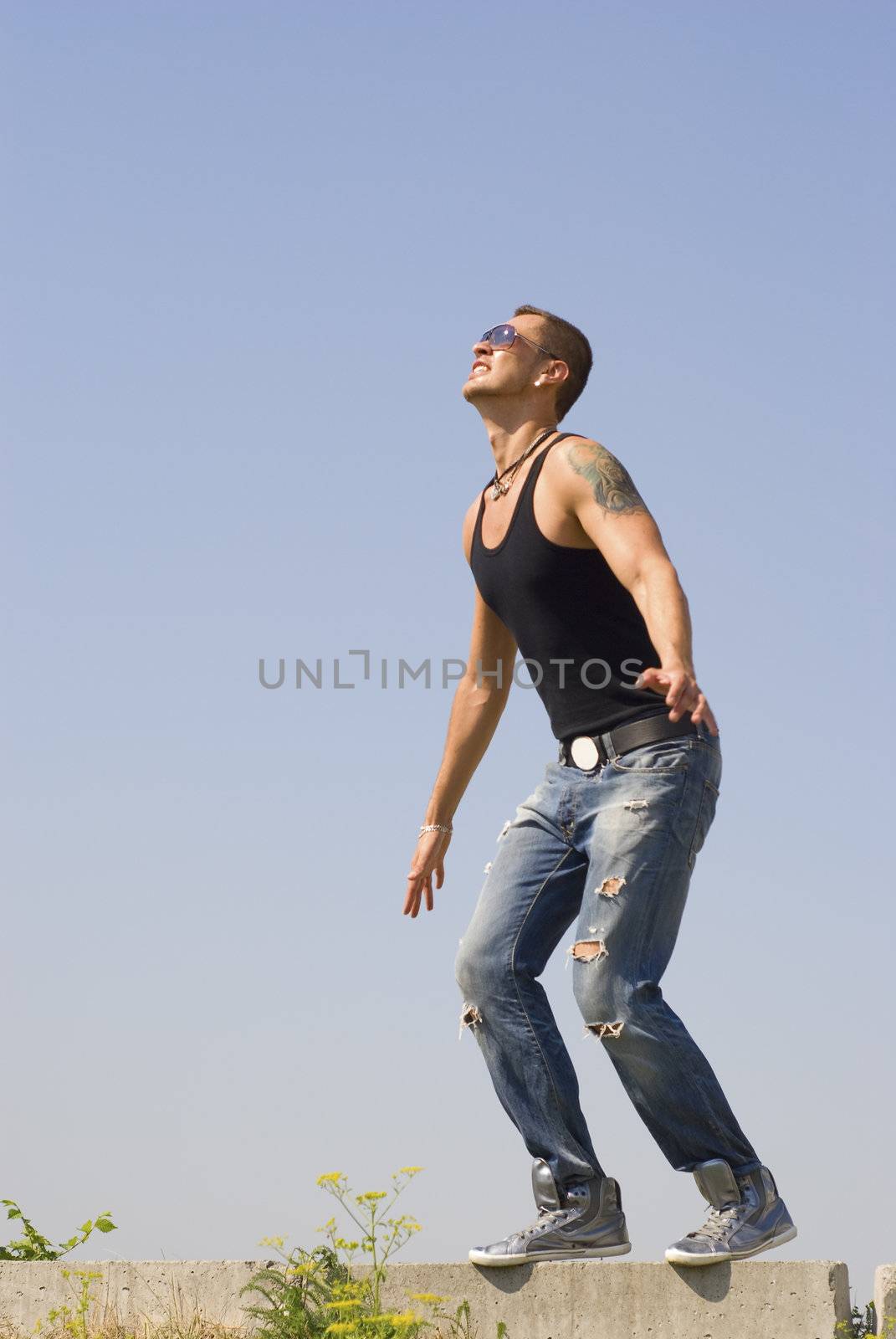 Expressive man strained pose with opend arms stays in protextive poze. He wears sun glasses and has tatoo on  the left shoulder. Tattoo represent the person in a hood.