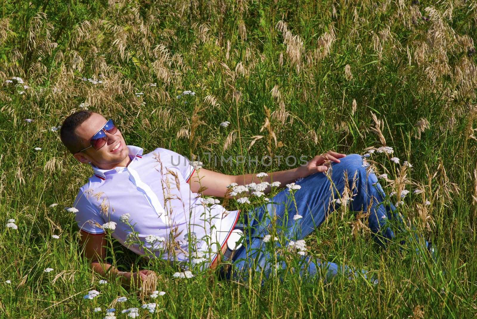 Young man in jeans with teeth smile is laying on summer field. Green grass surrounds him.
