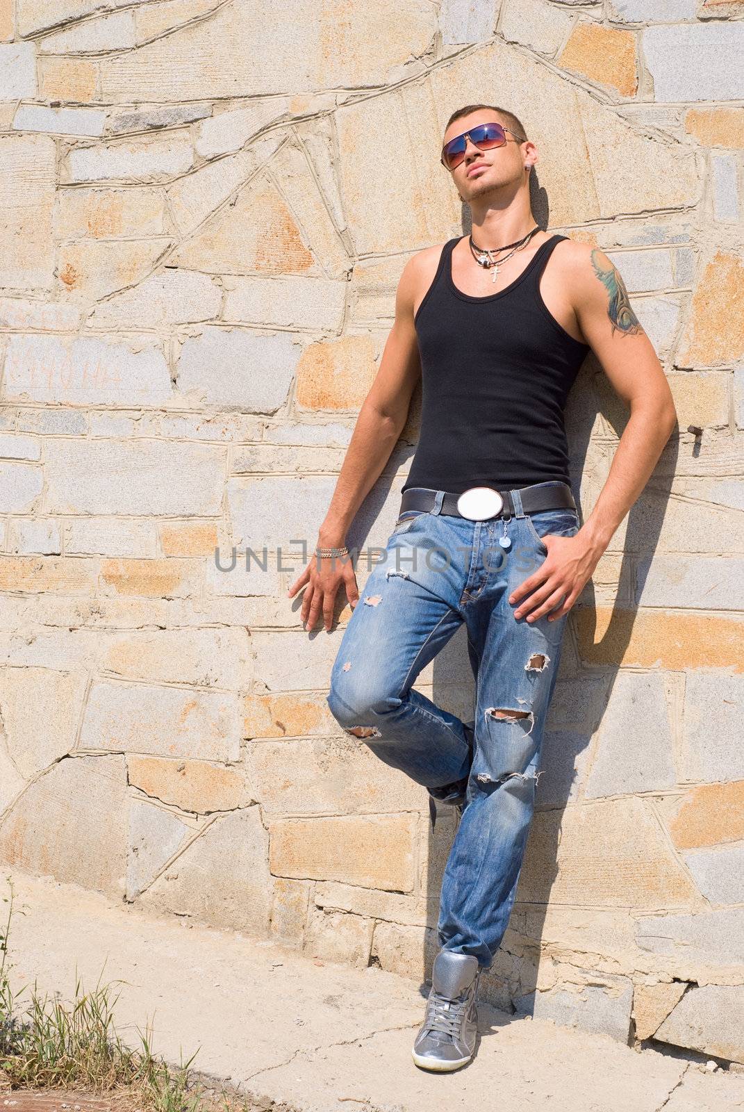 Person in sun glasses stands near the orange wall and looks at the sun. He has tattoo and earrings.