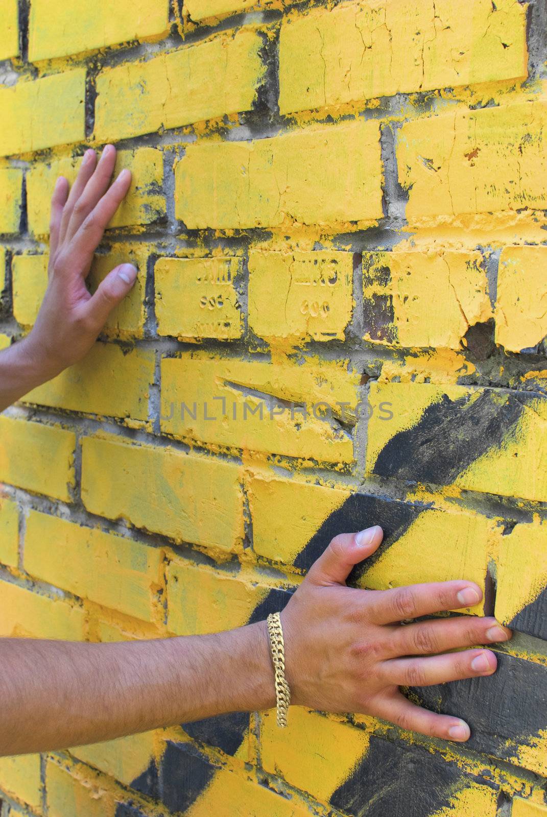 Hands of the young man are touching the wall. There is a yellow bracelet on right arm.