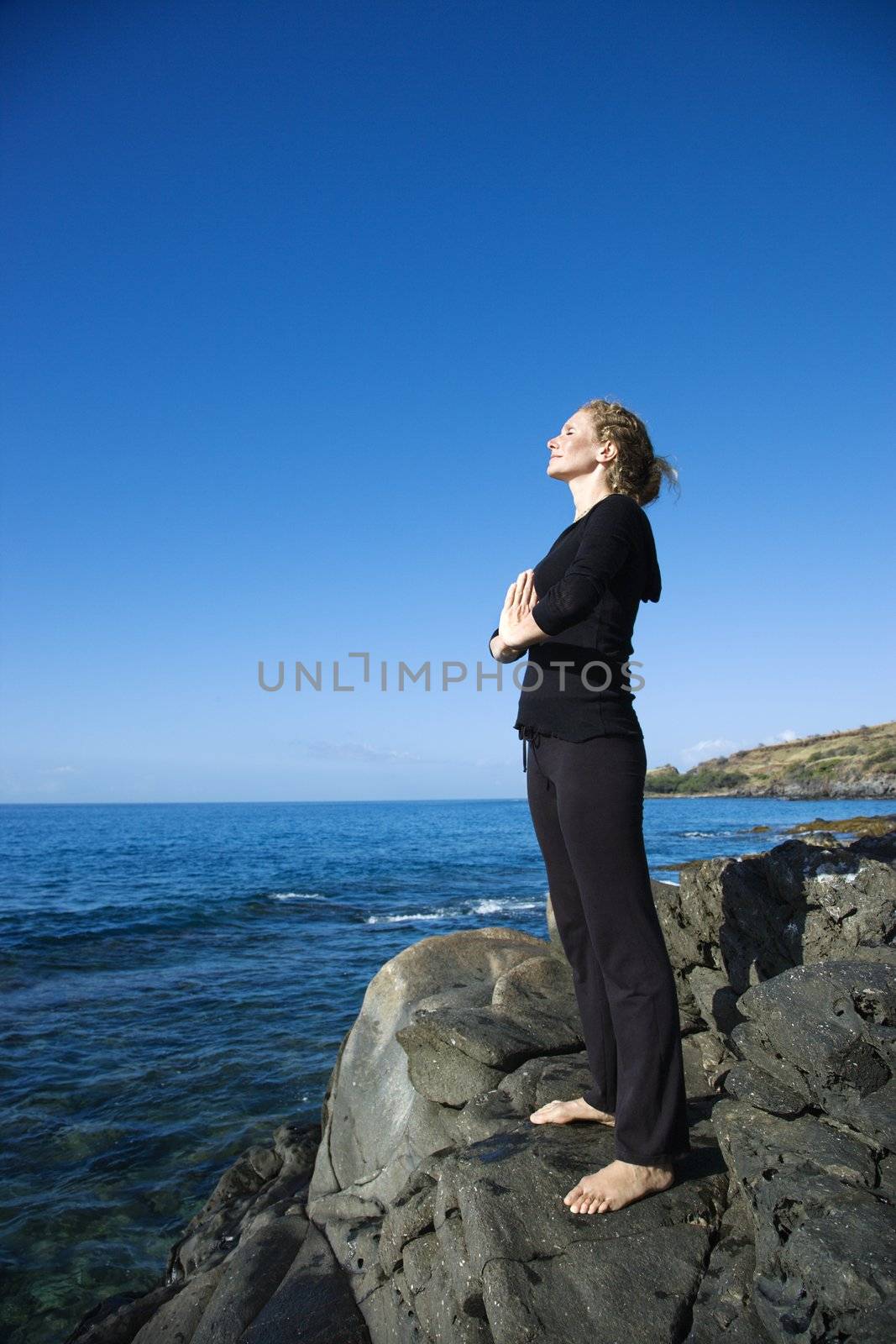 Caucasian mid-adult woman standing on rocky coast of Maui, Hawaii, meditating.