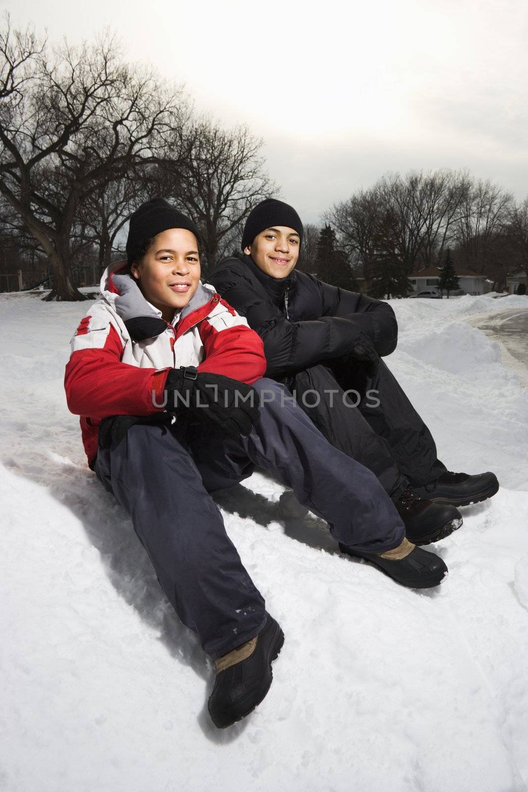 Two boys sitting in snow wearing coats and hats and smiling.