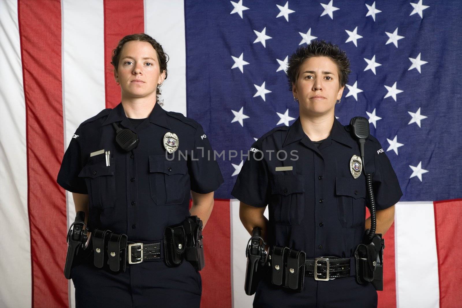 Portrait of two mid adult Caucasian policewomen standing with arms behind their backs and American flag as backdrop.