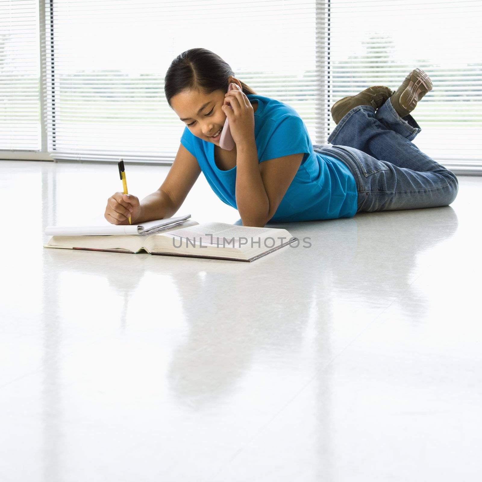 Asian preteen girl lying on floor doing homework while talking on cell phone.