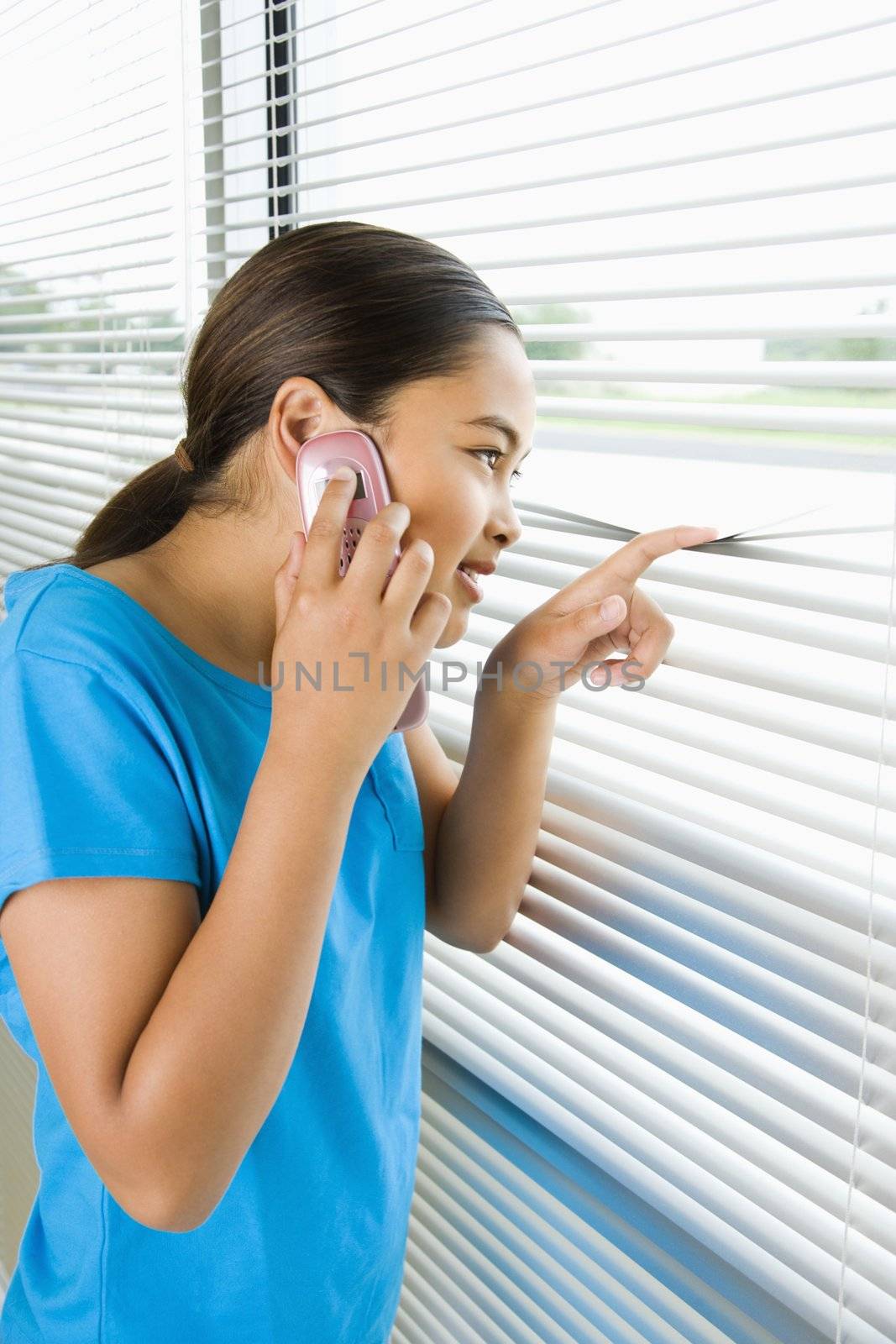 Side view of Asian preteen girl looking through blinds out window while talking on cell phone.