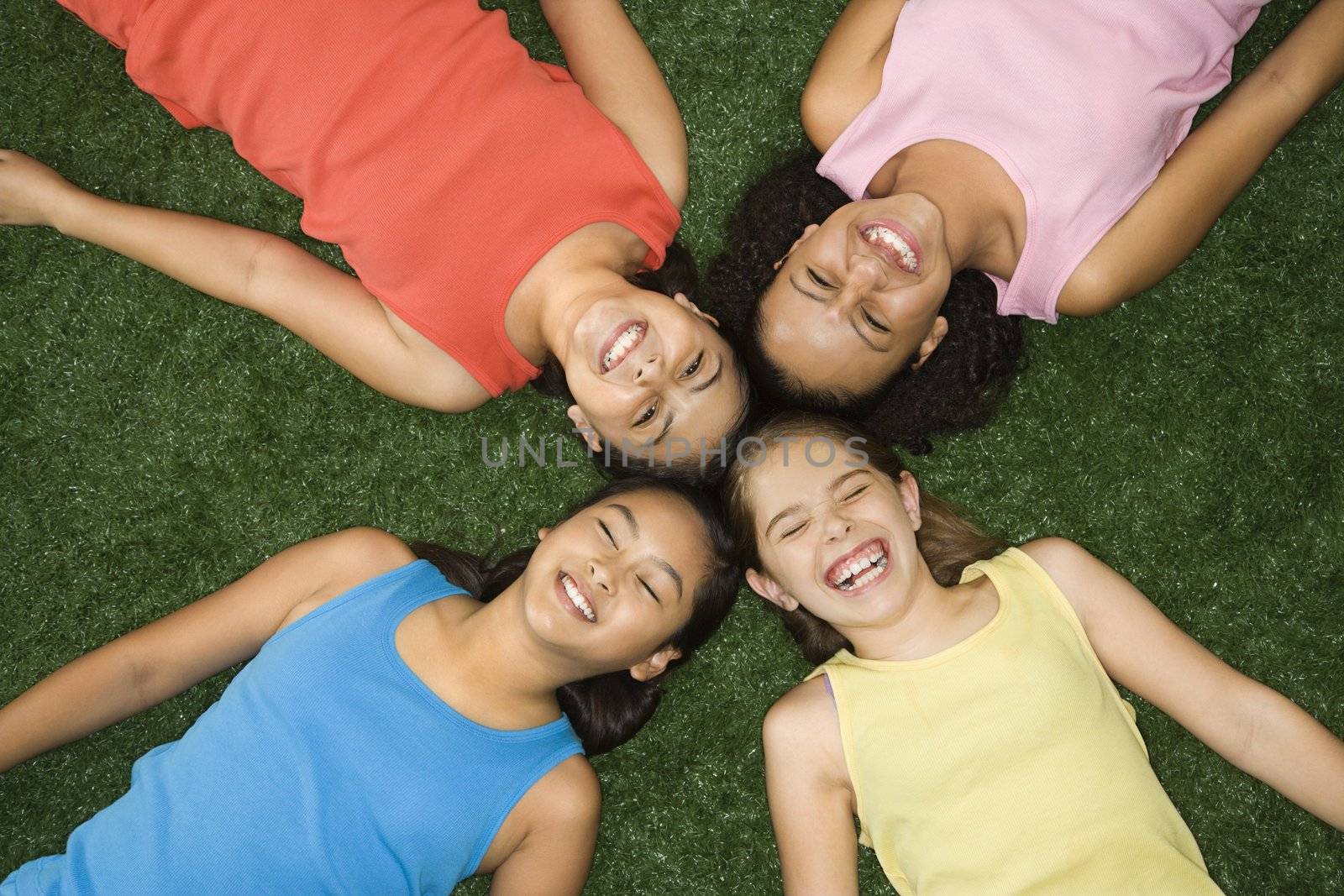 Above view of four girls lying on artificial grass with heads together laughing.