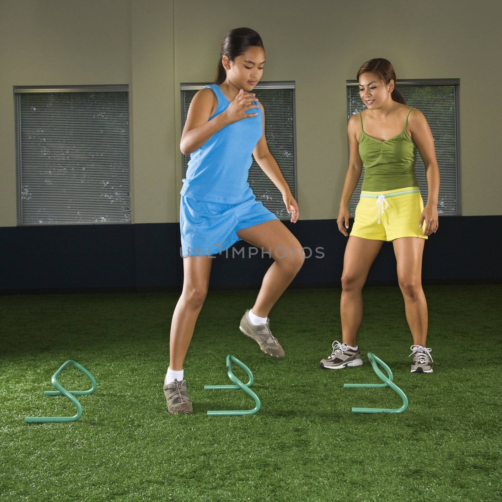 Girl jumping step hurdles in indoor gym while woman coach watches.