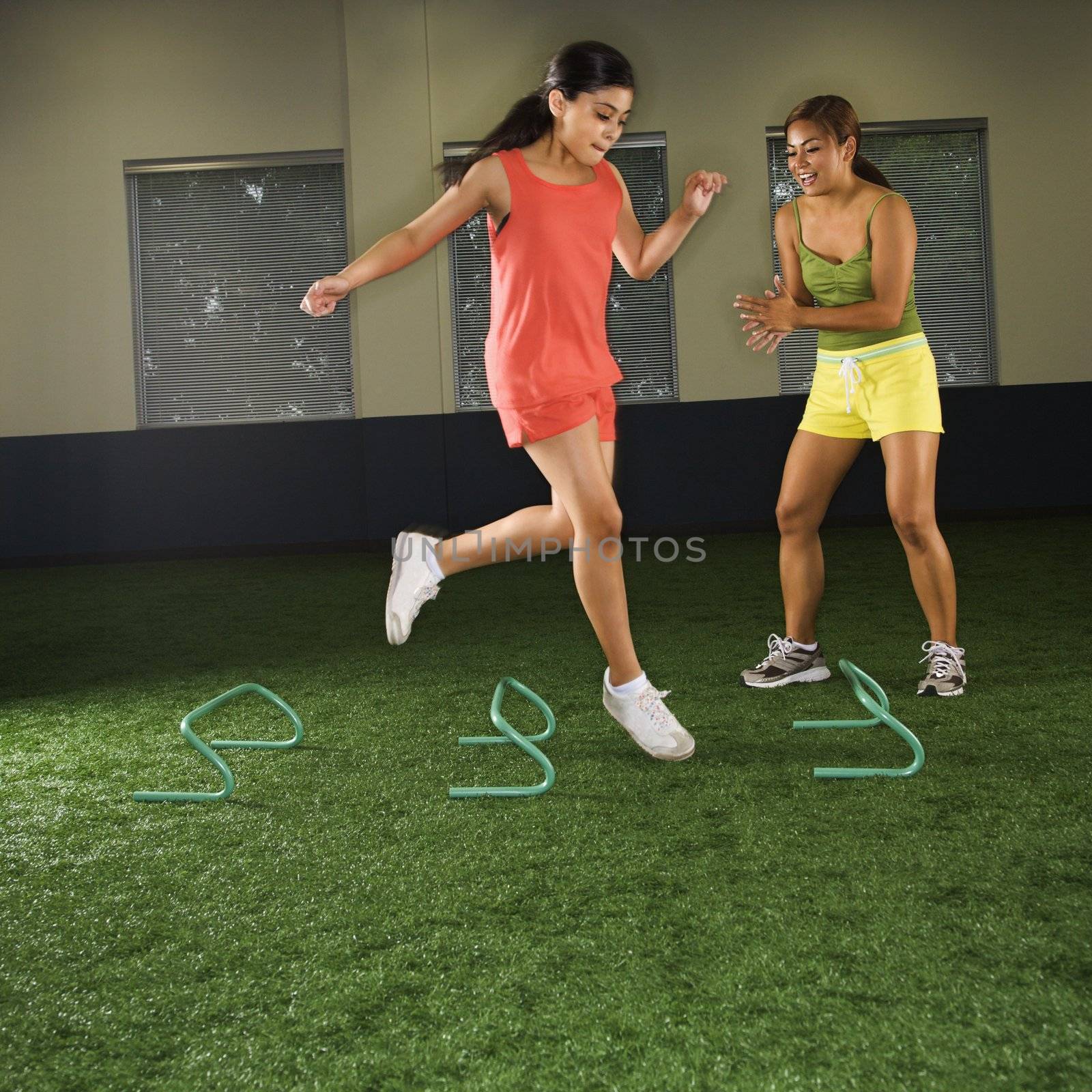 Girl jumping step hurdles in indoor gym while woman coach watches.