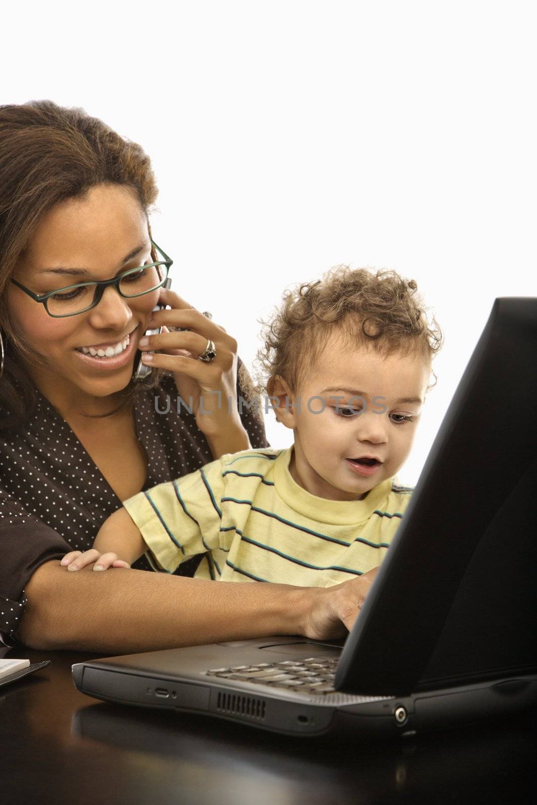 African American mid adult businesswoman at work on laptop and cell phone with toddler son on lap.