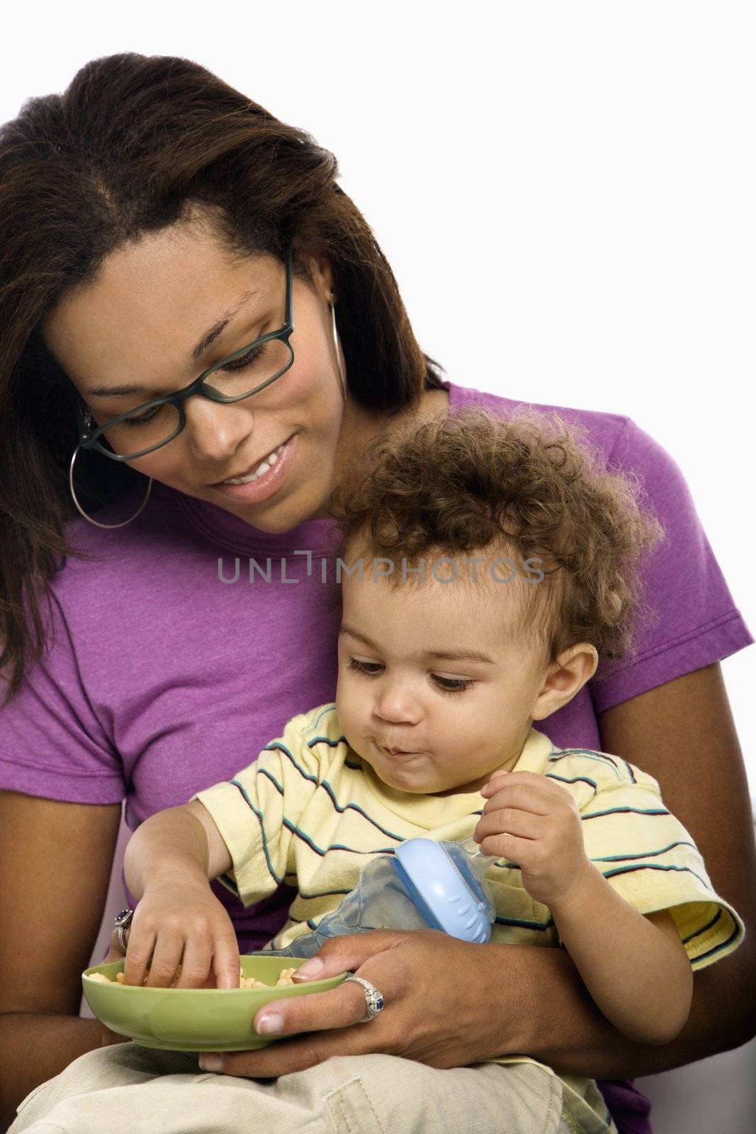 African American mid adult mom sitting with toddler son on lap eating cereal.