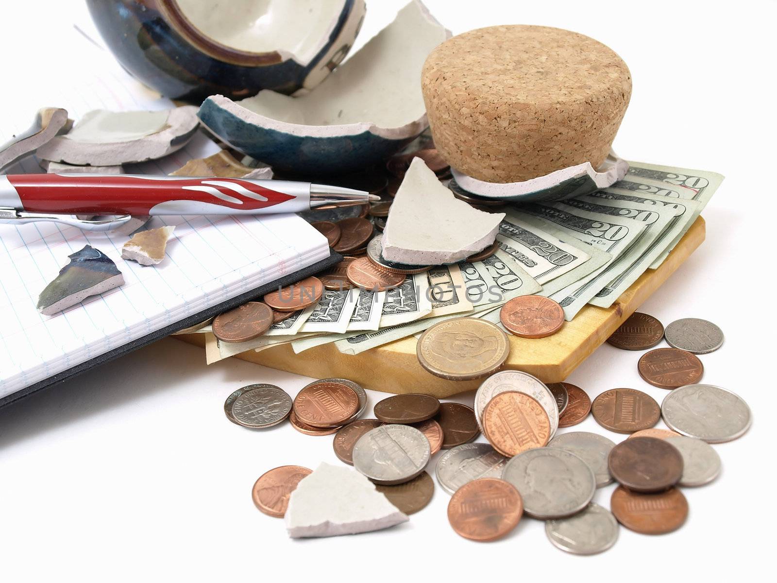 An open ledger and pen next to a broken ceramic bank on a pile of loose United States coins and currency.