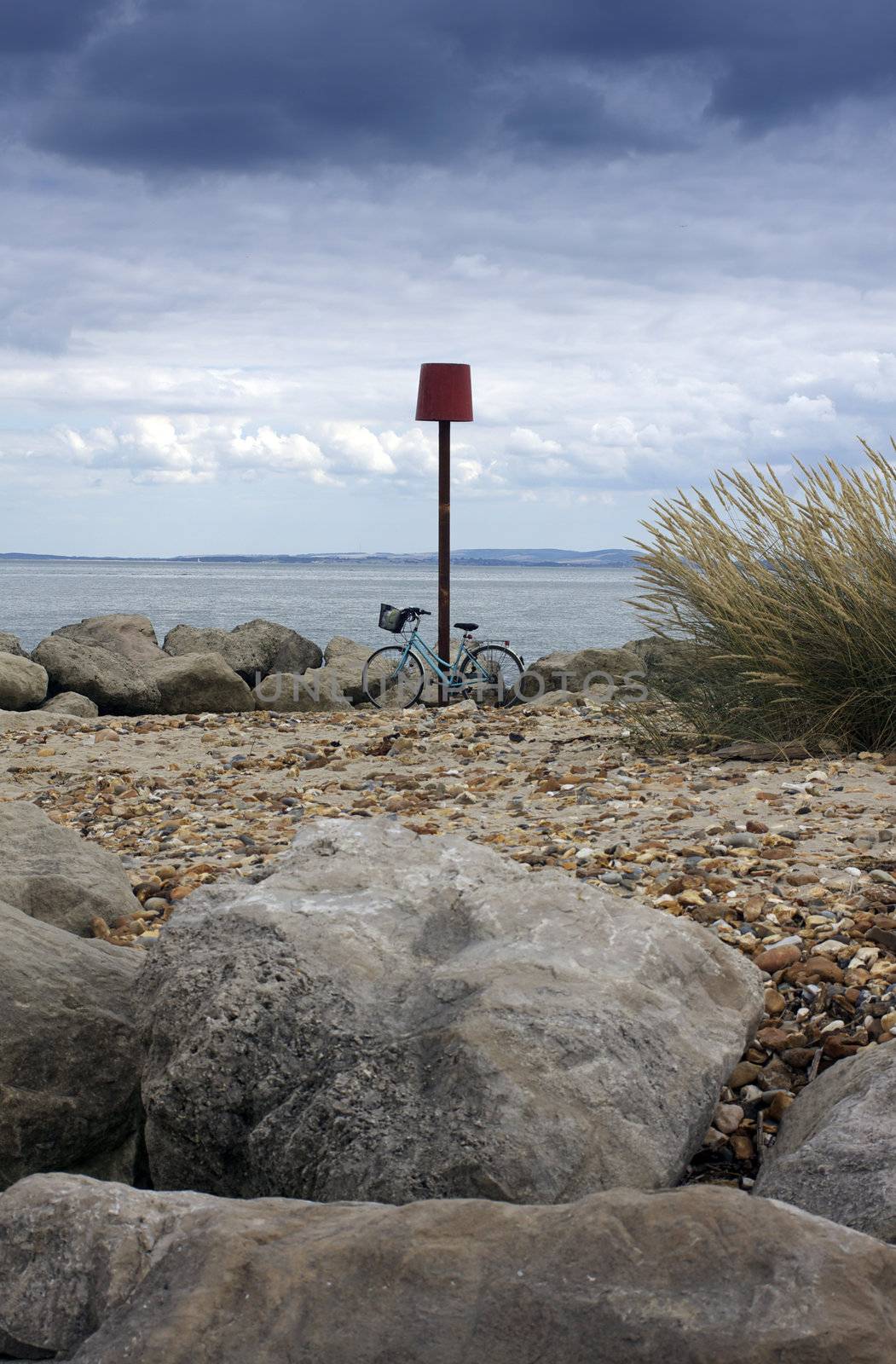 A bicycle on a beach setting. Bicycle propped up against a groyne beach marker. Coastal rocks and a pebbled beach to the foreground, a stormy landscape horizon of the isle of wight visible to the background. Location in Christchurch, Dorset, England. set on a portrait format.