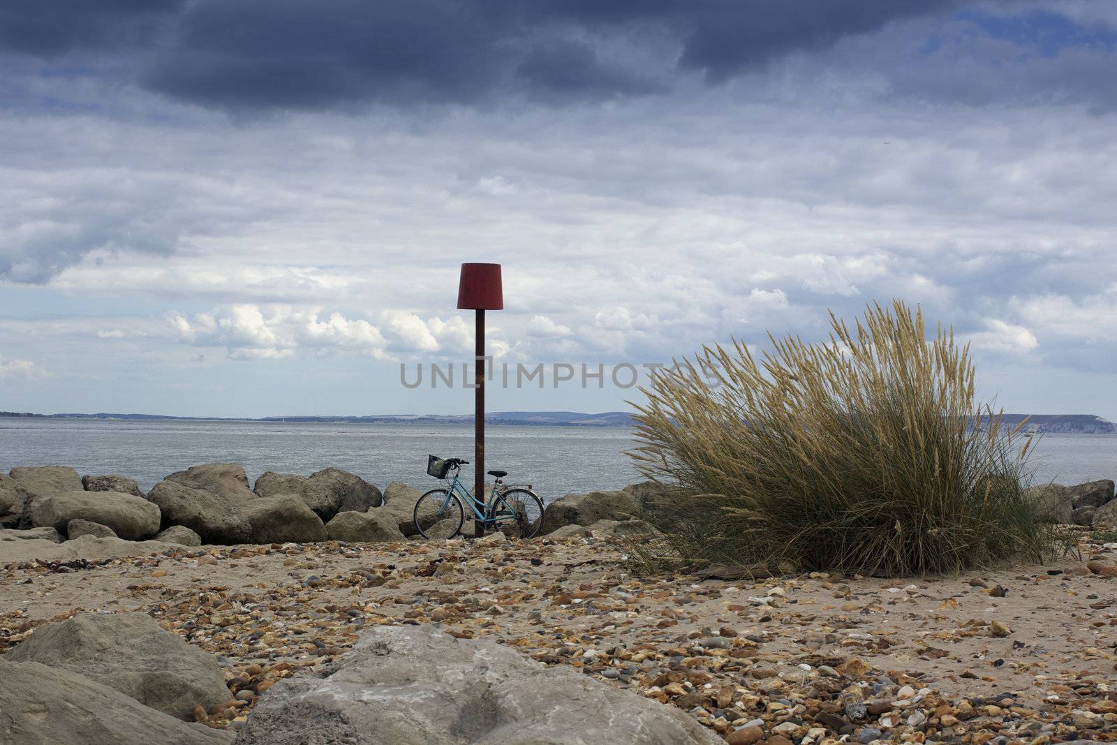 A bicycle on a beach setting. Bicycle propped up against a groyne beach marker. Coastal rocks and a pebbled beach to the foreground, a stormy landscape horizon of the isle of wight visible to the background. Location in Christchurch, Dorset, England. Set on a landscape format.