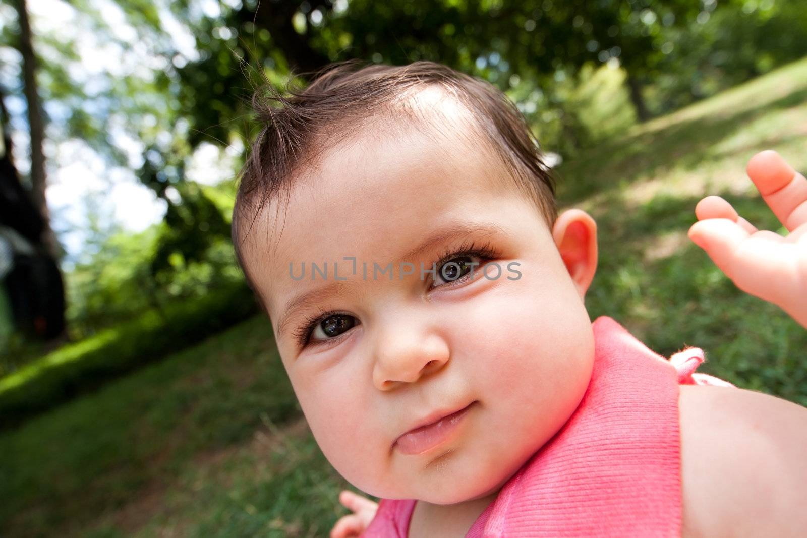 Caricature cute funny distorted baby face with big chubby cheeks of a Caucasian Hispanic infant girl with glossy eyes, in a park.