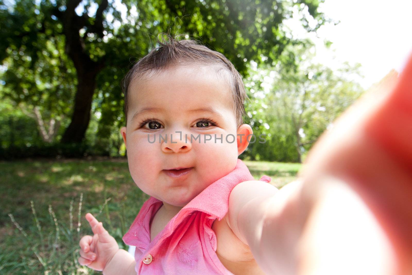 Cute funny happy baby infant sticking arm out as if she's taking self portrait photo in a park.